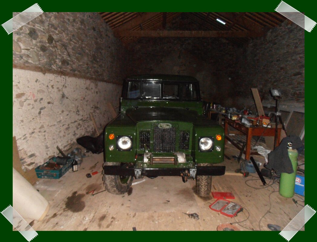 A green Land Rover Series 2 in a darkish barn with a bench full of tools next to it.
